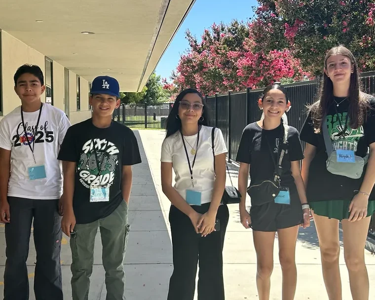 Five middle school students on a campus walkway smiling at the camera in their lanyards